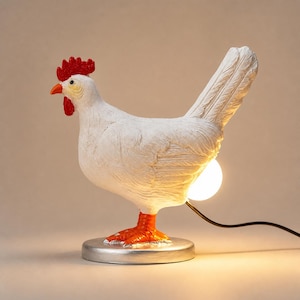 May include: A white chicken-shaped table lamp with a textured body, a bright red comb and wattle, and orange feet. The lamp stands on a circular silver base and features a glowing white light bulb positioned at the rear, connected by a black cord.