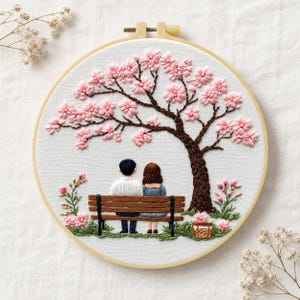 May include: A circular embroidery hoop containing a fabric panel with a hand-stitched scene of two figures sitting on a wooden bench under a large cherry blossom tree with pink flowers. A small woven basket sits on the green grass near the tree trunk.