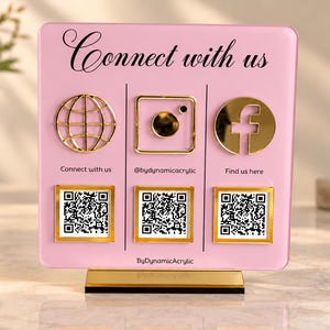 May include: A square light pink acrylic social media sign with a gold base. It features the text Connect with us, gold icons for a website, Instagram, and Facebook, and three QR codes with gold frames. The text @bydynamicacrylic is included.