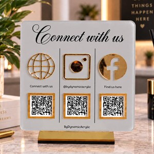 May include: A square white acrylic sign with gold accents featuring the text Connect with us, three gold social media icons, and three QR codes. Below the icons, the text reads Connect with us, @bydynamicacrylic, and Find us here.
