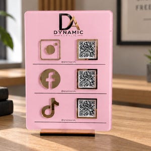 May include: A rectangular light pink acrylic sign featuring the gold DYNAMIC ACRYLIC logo at the top. It displays three social media icons—Instagram, Facebook, and TikTok—in gold, each paired with a square QR code and the handle @dynamicacrylic.