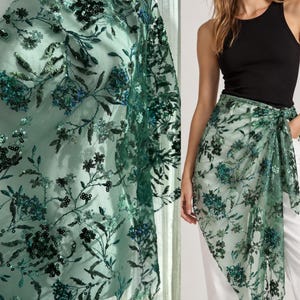 Op de afbeelding: Een doorschijnende, lichtgroene mesh sarong met een ingewikkeld bloemenpatroon geborduurd met donkergroene en zwarte pailletten. De stof is lichtgewicht en semi-transparant, met een gedetailleerd botanisch ontwerp over het gehele oppervlak.