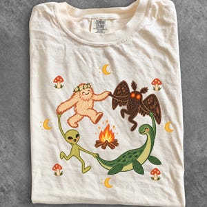 May include: A cream-coloured t-shirt featuring a graphic of a yeti, a moth-like creature, an alien, and a sea monster holding hands in a circle around a campfire. The design includes small red mushrooms and yellow crescent moons surrounding them.