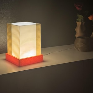 Può includere: Una lampada da tavolo quadrata con un corpo cubico bianco traslucido e luminoso con lati dai toni dorati. La lampada poggia su una base quadrata arancione vibrante e testurizzata e include un sottile cavo di alimentazione bianco sul retro.
