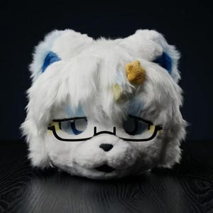 May include: A white plush animal head mask featuring long, shaggy faux fur, blue inner ear accents, and a small yellow star detail. The mask includes large, expressive eyes and a pair of black-rimmed glasses resting across the bridge of the nose.