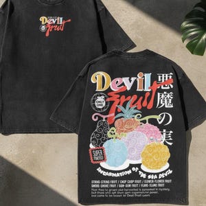 Könnte beinhalten: Ein schwarzes T-Shirt mit kurzen Ärmeln, das auf der Vorderseite ein kleines Motiv und auf der Rückseite ein großes, farbenfrohes Design zeigt. Die Rückseite zeigt verschiedene stilisierte Früchte, den Text "Devil Fruit", japanische Schriftzeichen und eine Liste von Fruchtnamen.