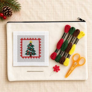 May include: A needlepoint kit featuring a square canvas with a stitched Christmas tree design, four skeins of embroidery floss in red, green, brown, and yellow, a metal needle, a red flower-shaped needle threader, and orange scissors.