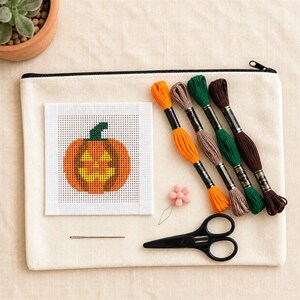 Puede incluir: Un kit de punto de cruz que incluye una tela blanca cuadrada con un diseño de calabaza, cuatro madejas de hilo de bordar en naranja, beige, verde y marrón, un imán para agujas con forma de flor, una aguja de coser y tijeras negras sobre un estuche.