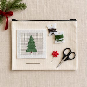 May include: A beige canvas zipper pouch featuring a cross-stitch pattern of a green Christmas tree on white fabric, two plastic thread bobbins with dark brown and green embroidery floss, a red flower-shaped needle minder, and metal scissors.