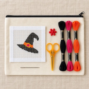 May include: A cream-colored canvas zipper pouch containing a needlepoint canvas featuring a black witch hat with an orange band, a single sewing needle, a red flower-shaped needle minder, orange embroidery scissors, and three skeins of embroidery floss in black, hot pink, and bright orange.