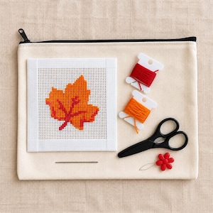 Puede incluir: Un estuche de lona beige con cremallera que contiene un cuadrado de punto de aguja con un diseño de hoja naranja y roja, dos bobinas de plástico blanco con hilo de bordar rojo y naranja, tijeras negras, una aguja de metal y un alfiler de flor roja.