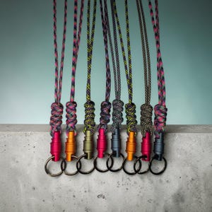 May include: Eight lanyards featuring multicoloured braided cords with intricate knotting details. Each cord is attached to a cylindrical metal connector in various colours including red, orange, olive, and dark grey, finished with a metal key ring.