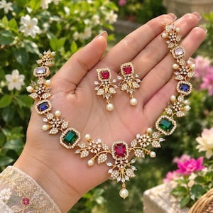 May include: A gold-toned necklace and earring set featuring rectangular-cut red, blue, and green gemstones. The jewelry is accented with clear white crystals and small white pearls arranged in floral-inspired patterns along the metal links.
