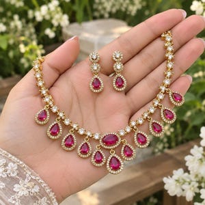 May include: A gold-toned jewelry set consisting of a necklace and matching drop earrings. The necklace features a series of teardrop-shaped fuchsia stones surrounded by clear white crystals, with a central oval fuchsia stone. The earrings mirror the design.