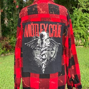 May include: A red and black buffalo plaid long-sleeve shirt with a bleached, distressed finish. A large black rectangular patch is sewn onto the back, featuring the text MÖTLEY CRÜE in red above a white graphic of a winged snake coiled around a sword.