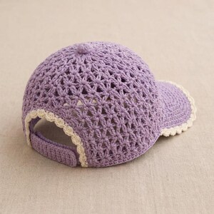 Pu&ograve; includere: Un berretto da baseball color lavanda realizzato in filato lavorato all'uncinetto con un motivo a stella traforato sulla corona. Il berretto include una visiera solida e un cinturino posteriore, entrambi rifiniti con un bordo smerlato color crema.