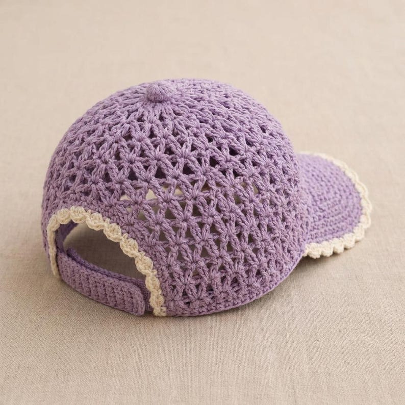 Pu&ograve; includere: Un berretto da baseball color lavanda realizzato in filato lavorato all'uncinetto con un motivo a stella traforato sulla corona. Il berretto include una visiera solida e un cinturino posteriore, entrambi rifiniti con un bordo smerlato color crema.