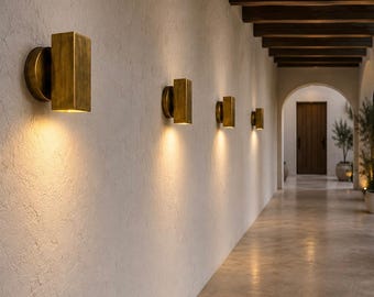 May include: A series of rectangular wall sconces with a brushed brass finish mounted on a textured off-white wall. Each fixture features a circular base and emits a warm, downward-facing light that illuminates the wall surface below the lamp.