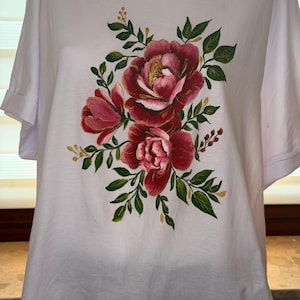 Könnte beinhalten: Ein weißes T-Shirt mit kurzen Ärmeln und einem handgemalten Blumenmotiv auf der Vorderseite. Das Design zeigt große, geschichtete Pfingstrosen in dunklen Rot-, Magenta- und Rosatönen, akzentuiert durch grüne Blätter und kleine goldfarbene Details.