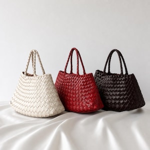 Op de afbeelding: Drie gevlochten handtassen in crème, rood en donkerbruin. Elke tas heeft een gestructureerd, uitlopend silhouet met een opvallend vlechtpatroon over het hele lichaam en twee bijpassende gevlochten handvatten om de tas te dragen.