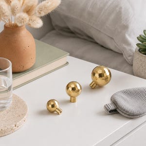 May include: Three spherical cabinet knobs in a polished gold finish, each featuring a faceted, hammered texture. The knobs are arranged in a row, showing a graduated size progression from smallest to largest, with a cylindrical base for mounting.