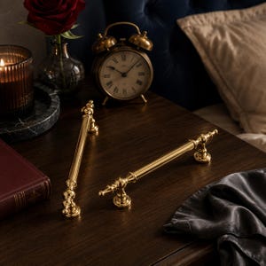 May include: Two polished gold-tone metal cabinet pulls with a cylindrical bar design and decorative finials at each end. The hardware features a smooth, reflective finish and circular mounting bases, presented as a pair of matching furniture handles.