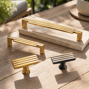Puede incluir: Una colección de cuatro herrajes para muebles que incluye dos tiradores rectangulares largos en acabado dorado cepillado y dos pomos rectangulares más pequeños, uno en acabado dorado cepillado y otro en negro mate, todos con ranuras horizontales.