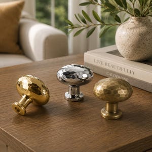 May include: Three cabinet knobs with a rounded, hammered texture. One knob is polished gold, one is polished silver, and one is a brushed antique brass finish. Each knob features a circular base and a smooth, reflective, dimpled surface design.