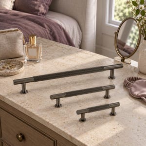 May include: Three dark metallic cabinet pulls of varying lengths, each featuring a cylindrical bar with a textured, ribbed center section and smooth ends, supported by two vertical posts that attach to the mounting surface.