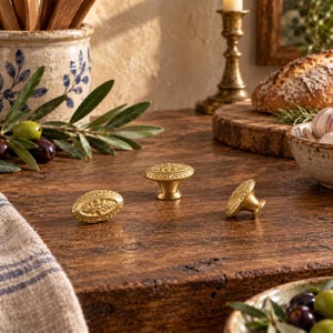 May include: Three round brass cabinet knobs with a textured, decorative edge and a central star-like motif. The knobs have a smooth, metallic finish and a tapered base, displayed on a dark wooden surface with a warm, golden-toned appearance.