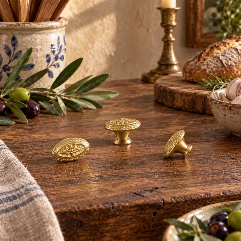 May include: Three round brass cabinet knobs with a textured, decorative edge and a central star-like motif. The knobs have a smooth, metallic finish and a tapered base, displayed on a dark wooden surface with a warm, golden-toned appearance.