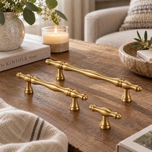 May include: Three brass-toned metal cabinet pulls and a knob with a smooth, curved design. The hardware features a central elongated body that tapers toward the ends, supported by two cylindrical posts, while the knob has a single central post.