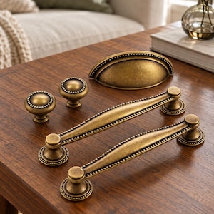 May include: A collection of antique brass hardware including two long arched drawer pulls, two round cabinet knobs, and one semi-circular cup pull. Each piece features a beaded edge detail and a weathered, metallic finish.