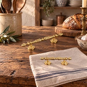May include: Two gold-toned metal cabinet pulls featuring an ornate, textured design with decorative finials at each end. The pulls have circular mounting bases with intricate floral-like patterns and a central bar with detailed engravings.
