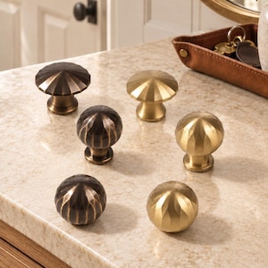 May include: A collection of six metal cabinet knobs arranged in two columns. The left column features three dark bronze knobs with a faceted, ribbed design. The right column displays three brushed gold knobs with a matching faceted, ribbed shape.
