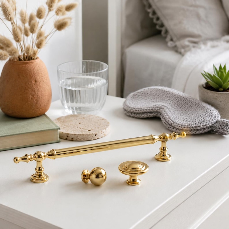 May include: A set of three polished gold-tone metal cabinet hardware pieces, including one long horizontal bar handle with decorative ends and two smaller round knobs, all featuring a smooth, reflective finish and classic cylindrical shapes.