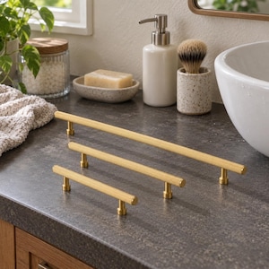 May include: Three gold-toned metal cabinet pulls with a textured, knurled surface. Each pull features a long, cylindrical bar supported by two small, circular mounting posts. The pulls are arranged in a row, showing three different lengths.