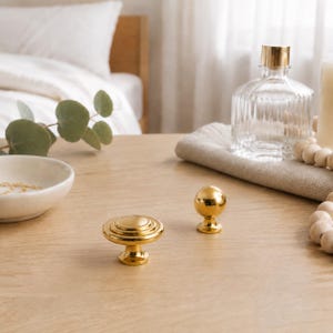 May include: Two gold-toned metal cabinet knobs are displayed. One knob features a flat, circular, tiered design with concentric rings, while the other is a smooth, spherical ball shape. Both knobs have a polished, reflective finish.