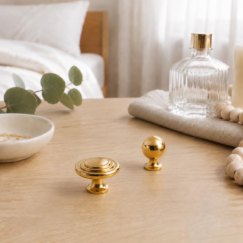 May include: Two gold-toned metal cabinet knobs are displayed. One knob features a flat, circular, tiered design with concentric rings, while the other is a smooth, spherical ball shape. Both knobs have a polished, reflective finish.