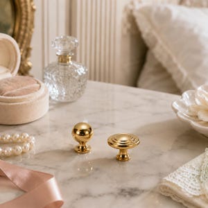 May include: Two polished gold-toned metal cabinet knobs. One knob features a smooth spherical head, while the other has a flat, circular top with concentric ridged detailing. Both knobs have a tapered base and a reflective, metallic finish.