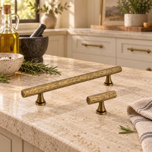 May include: A long cabinet pull and a smaller matching T-shaped knob, both featuring a textured, hammered antique brass finish. Each piece has a cylindrical bar design supported by conical bases, showcasing a consistent metallic aesthetic.