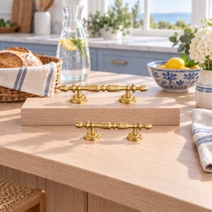 May include: Two ornate gold-toned metal cabinet pulls featuring intricate leaf-like detailing along the central bar and decorative circular bases. The hardware is displayed on a light-colored wooden surface, showcasing a polished finish and a classic, symmetrical design with two mounting points on each handle.