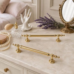 May include: Two polished gold-tone metal cabinet pulls featuring a cylindrical bar design with decorative finials at each end. The hardware has a smooth, reflective finish and includes two circular mounting bases for secure attachment to furniture.