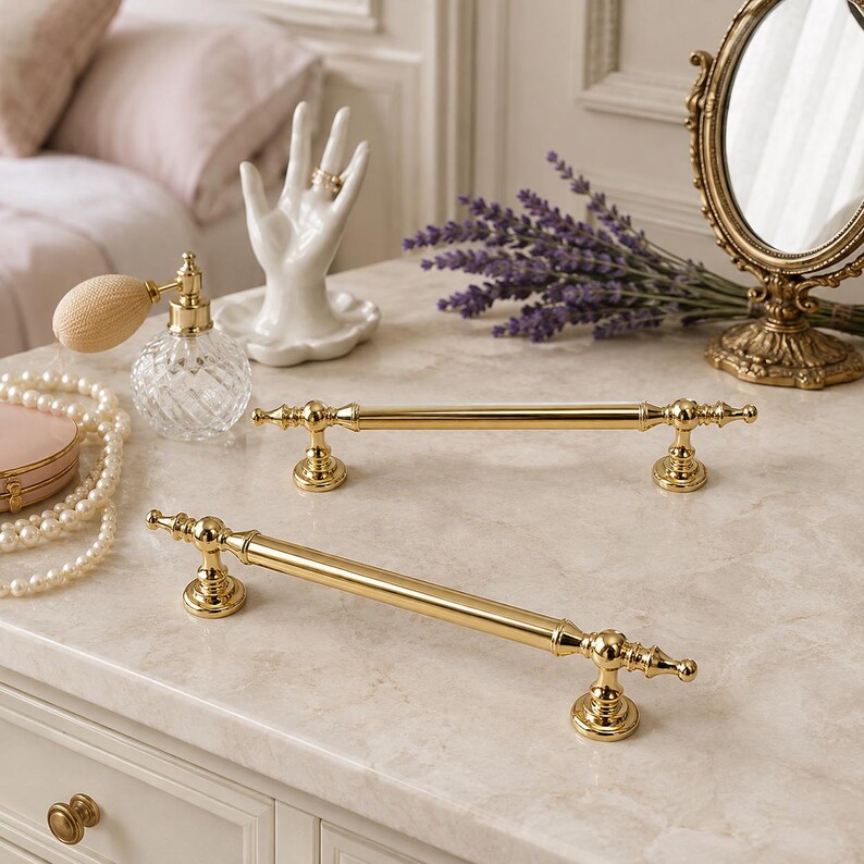 May include: Two polished gold-tone metal cabinet pulls featuring a cylindrical bar design with decorative finials at each end. The hardware has a smooth, reflective finish and includes two circular mounting bases for secure attachment to furniture.