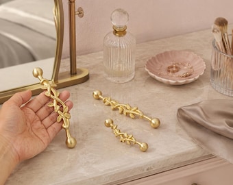 May include: Three gold-toned metal cabinet pulls featuring a vine and leaf design with spherical ends. The pulls are finished in a matte gold texture, showcasing a botanical motif that wraps around the central bar of each handle.