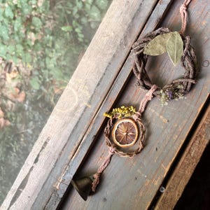 May include: A hanging decoration featuring two interconnected brown twig wreaths suspended by pink twine. The top wreath is adorned with two green leaves and small dried flowers. The bottom wreath holds a dried orange slice, yellow sprigs, and a metal bell.