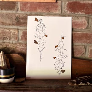May include: A rectangular white print featuring two vertical black ink illustrations of flowering plant stalks. Several small, orange and brown patterned butterflies are positioned on and around the stems of the plants, creating a delicate nature-inspired composition.
