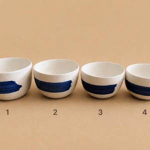 Peut inclure: Quatre bols en céramique blanche disposés en rangée, chacun présentant un motif de coup de pinceau bleu foncé horizontal. Les bols sont numérotés de un à quatre en texte brun sous chaque article, montrant une légère variation de taille.