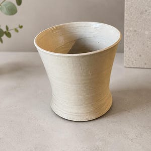 Peut inclure: Un récipient en céramique tourné à la main avec un bord évasé et une base effilée. L'extérieur présente un vernis blanc cassé lisse avec de subtiles stries horizontales et une texture visible. L'intérieur est creux avec une large ouverture.