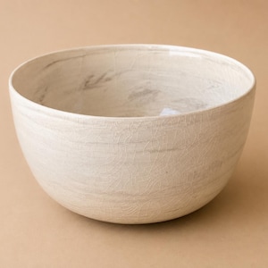 Peut inclure: Un bol rond en céramique avec un vernis de couleur crème présentant une texture fine et craquelée subtile, ainsi que de légères stries horizontales gris-brun qui enveloppent les surfaces intérieure et extérieure, créant un aspect organique.